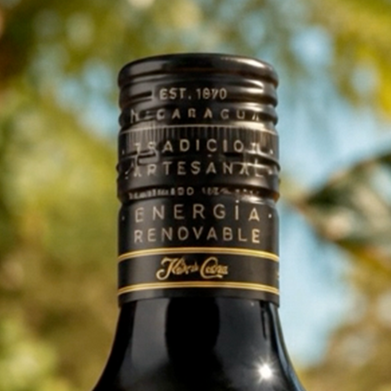 Close-up of a dark glass bottle neck with a black label reading 'ENERGIA RENOVABLE' and 'Kiber Coca' on a blurred outdoor background.