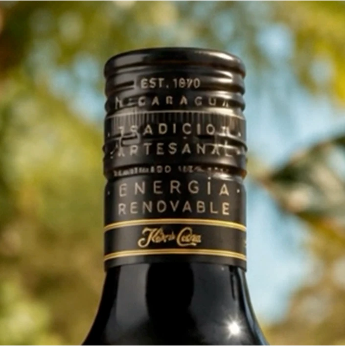 Close-up of a dark glass bottle neck with a black label reading 'ENERGIA RENOVABLE' and 'Kiber Coca' on a blurred outdoor background.