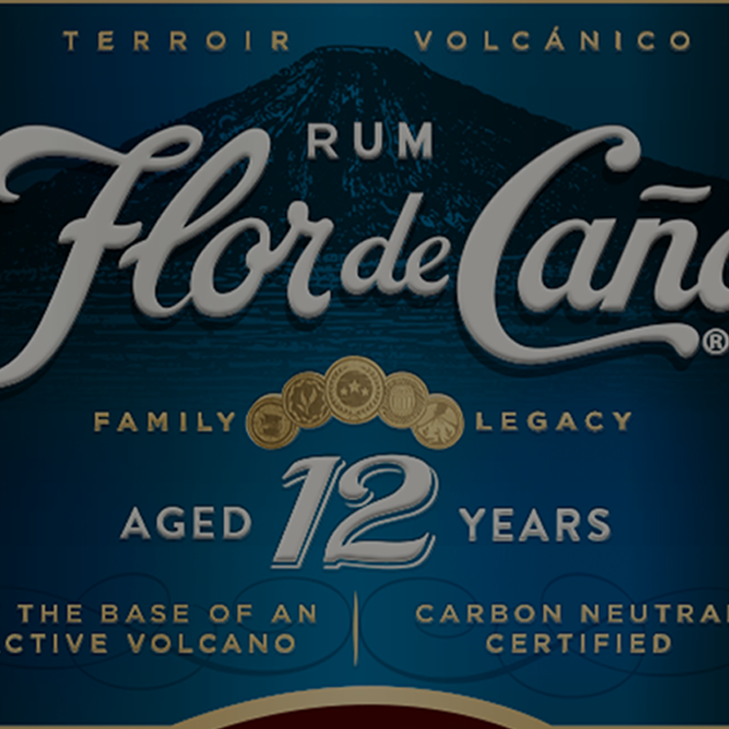 Rum Flor de Caño Terroir Volcánico aged 12 years rum bottle label design, featuring a dark blue background with a volcano graphic, white script logo, and gold accents.