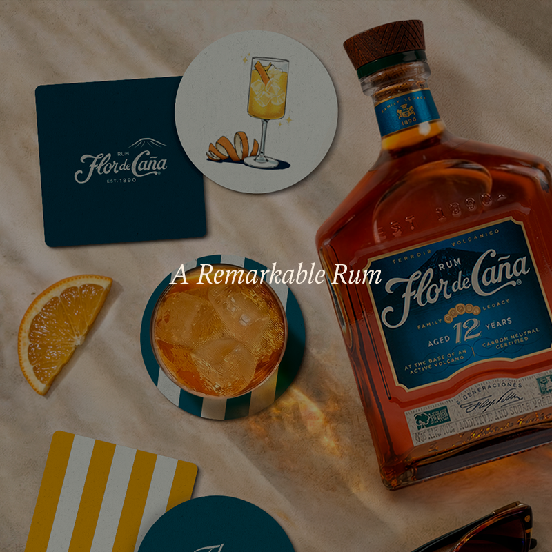 A bottle of Flor de Cána rum and matching branded coasters and drinkware arranged on a beige surface, with an orange slice and a cocktail glass in the scene. The image includes the text 'A Remarkable Rum' across the center.