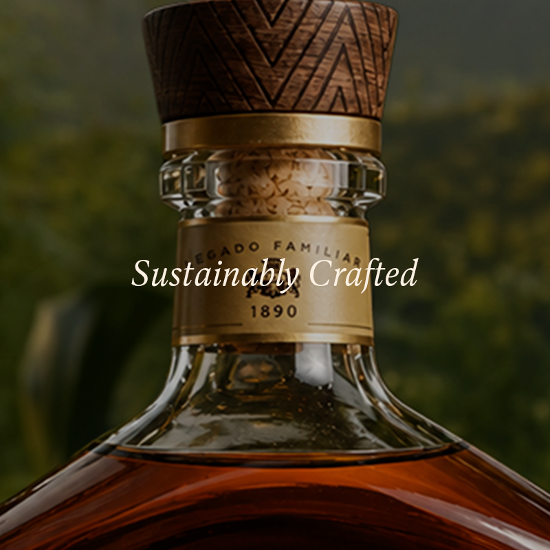 Close-up of a premium liquor bottle with amber liquid, a gold neck label reading 1890, a carved dark wood stopper, and the text 'Sustainably Crafted' overlaid across the center.