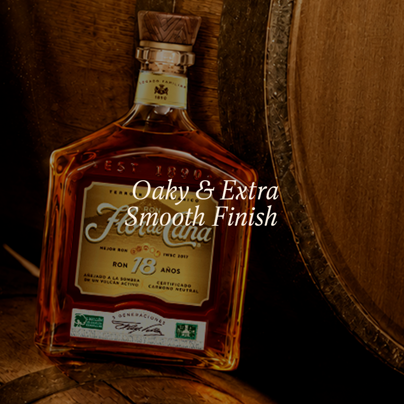 A bottle of Flor de Caña Ron 18 Años whiskey with a cork stopper, resting against wooden barrels. The image features a text overlay that reads 'Oaky & Extra Smooth Finish' in white script.