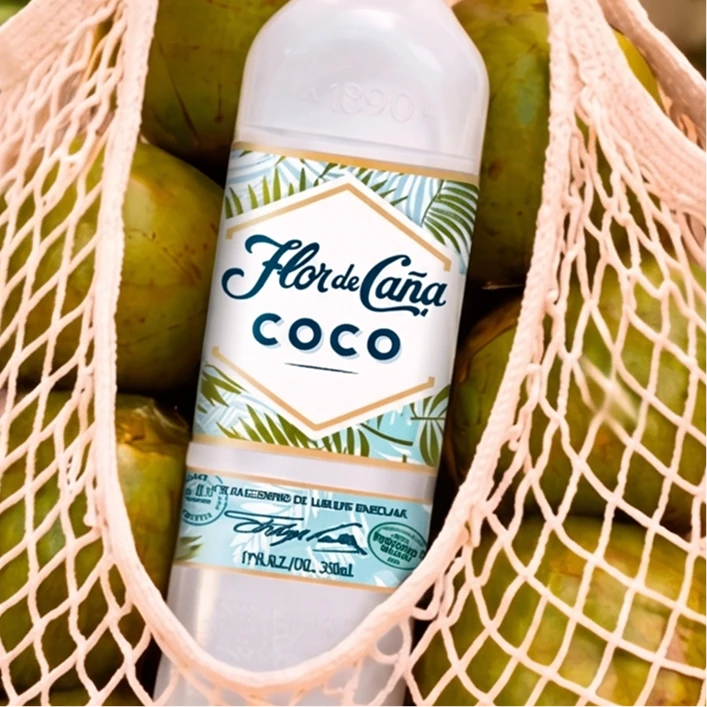 A bottle of Flor de Caña Coco liqueur placed inside a peach mesh bag filled with green coconuts. The bottle label features tropical palm leaves and the brand name 'Flor de Caña' with 'COCO' written below.